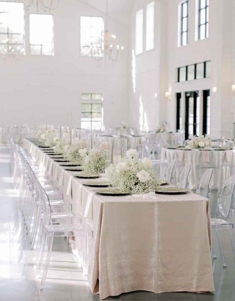 Long banquet table with elegant wedding decor rentals Houston featuring white florals, candles, and clear ghost chairs.